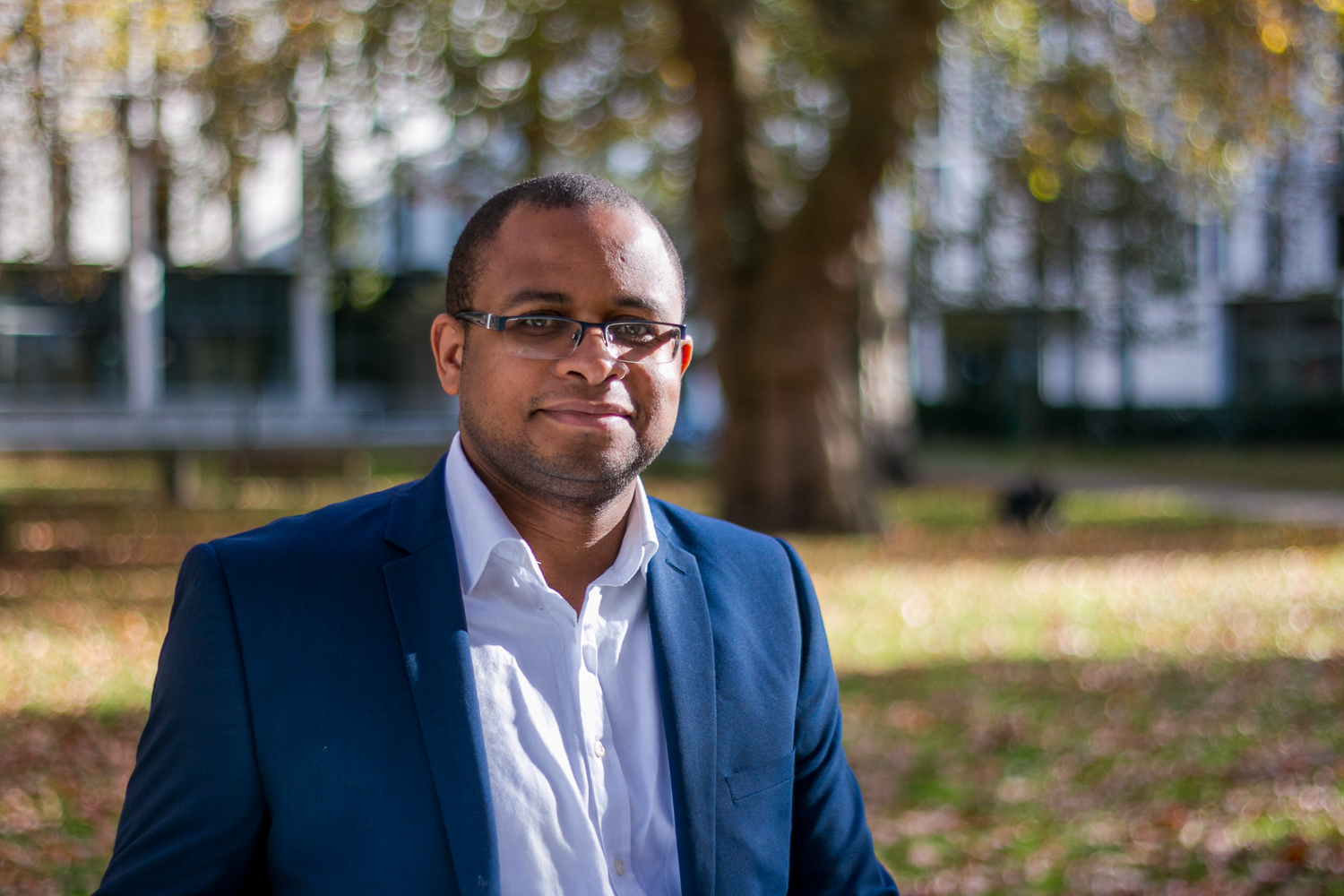 Photo of man dressed smartly, standing outside in a leafy green area, the sun is shining on him. He is slightly smiling